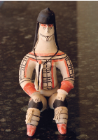 Imagem: Fotografia. Um boneco sentado, de pele clara, com pinturas no corpo em laranja, preta e sobre a cabeça, capacete de cor marrom-escuro. Ele está com as mãos sobre os joelhos.   Fim da imagem.