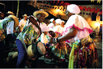 Imagem: Fotografia. À frente, um homem à esquerda de pele negra, com chapéu sobre a cabeça de cor bege, com camiseta de cor branca, com folhas verdes, parte inferior calça em azul. Sobre ele, pendurado, um tambor redondo pequeno de cor bege e parte em preto. À direita, uma mulher de pele negra, com lenço branco sobre a cabeça, com blusa branca com babado de cor branca, na parte inferior, borda em rosa. Na parte inferior, saia colorida em amarelo com estampas. Ao fundo, local com outras mulheres como mesmo tipo de roupa e homens. No alto, pendurado, bandeirinhas em amarelo, e vermelho e céu noturno preto, com luzes amarelas.  Fim da imagem.