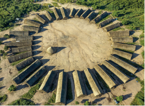 Imagem: Fotografia. Vista do alto de local com tocas grandes com o formato fino e telhado de triangular na cor bege-claro, formando um círculo. À esquerda, uma toca menor arredondada. Ao redor, grama de cor verde.  Fim da imagem.