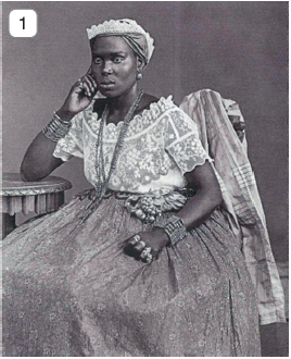 Imagem: Fotografia 1 em preto e branco. Mulher negra sentada em cadeira, com lenço sobre a cabeça, blusa de mangas curtas, claros e saia longa até os pés. Ela tem várias cores, pulseiras e anéis no dedo. Ela está com o cotovelo encostado em uma base redonda à esquerda, com mão debaixo de um queixo.  Fim da imagem.