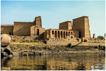 Imagem: Fotografia. À frente, rio com água escura e ao fundo, construção antiga, grande com forma retangular, aberturas na vertical com partes na vertical. No alto, céu em azul-claro.  Fim da imagem.