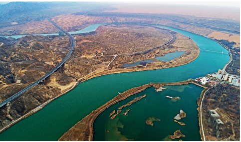 Imagem: Fotografia. Vista do alto de local com rio grande de cor verde com solo marrom à esquerda, rodovia longa de cor verde e mais à direita e na borda, rochas cinzas, local com solo marrom, algumas construções.  Fim da imagem.