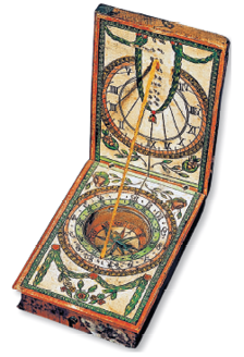 Imagem: Fotografia. Uma bússola com o formato arredondado ao centro, com rosa dos ventos. Na parte superior, base na vertical de cor branca, com ilustração ornadas em marrom e verde. Da parte de baixo para cima, na diagonal, com tampa na vertical. Fim da imagem.