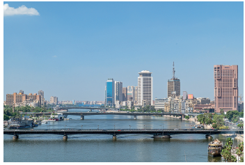 Imagem: Fotografia. Vista geral de local com rio de água escura, à frente uma ponte grande por onde passa automóveis e mais ao fundo, outras duas pontes. À esquerda e à direita, prédios de tamanhos pequenos, médios e grandes. À direita, prédio grande de cor marrom, outro de cor marrom com antena no alto, fina. Mais ao lado, prédio branco e outro em azul, espelhado. No alto, céu em azul-claro com vista parcial de nuvem à esquerda, em branco.  Fim da imagem.