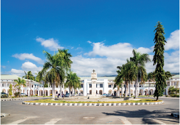 Imagem: Fotografia. Uma rua de cor cinza, com uma parte arredondada com vegetação verde, árvores de folhas verdes e em segundo plano, um casarão grande com dois andares, paredes de cor branca, com torre ao centro, com cruz na parte superior. À frente, local com arcos de cor branca. No alto, céu de cor azul-claro e nuvens brancas.  Fim da imagem.