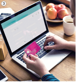 Imagem: Fotografia 3. Vista do alto, sobre mesa de madeira de cor marrom, com um notebook de cor cinza, com teclado de cor preta. A pessoa vista parcial de pele clara, segurando um cartão de cor rosa-escuro. À direita, um prato de cor branca com maçãs e um copo branco.  Fim da imagem.