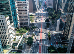 Imagem: Fotografia. Vista do alto de rua ao centro, de cor cinza e faixa ao centro, com automóveis passando, com algumas árvores de folhas verdes. À esquerda e à direita, prédios de tamanho médio e outros altos, espelhados.  Fim da imagem.