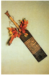 Imagem: Fotografia. Um colar com haste na vertical de cor bege, com penas coloridas na parte inferior de cor laranja. Mais abaixo, placa na vertical de cor marrom-escuro e ornamentos dentro de parte ao centro de cor bege.  Fim da imagem.