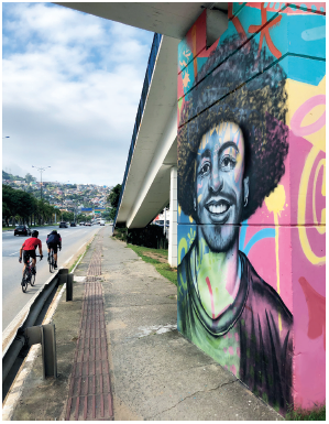 Imagem: Fotografia. À esquerda, vista parcial de rua de cor cinza, com dois ciclistas passando para frente. À direita, calçada de cor cinza, uma parede com ilustração de um homem com camiseta, cabelos encaracolados escuros, sorrindo e olhando para frente. Em segundo plano, partes com árvores e cidade com casas. No alto, céu azul-claro com nuvens brancas.  Fim da imagem.