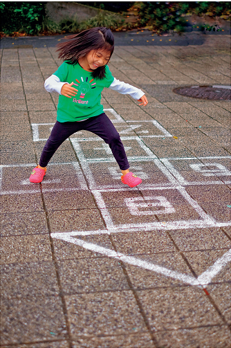 Imagem: Fotografia. Uma menina com cabelos lisos, olhos puxados, usando uma blusa de manga comprida, calça azul e sapatos rosas. Ela está saltando sobre uma amarelinha no chão e sorrindo. Fim da imagem.