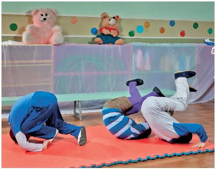 Imagem: Fotografia. Três crianças com as costas sobre um tatame com as pernas para cima. Fim da imagem.