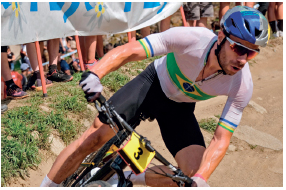 Imagem: Fotografia. Um homem com camiseta, short, capacete, óculos de sol, luvas e tênis. Ele está em cima de uma bicicleta. Fim da imagem.