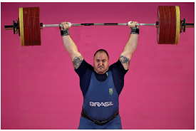 Imagem: Fotografia. Um homem robusto, com camiseta, macacão e um cinto na cintura. Ele está com os braços estendidos para cima segurando uma barra com ferro. Fim da imagem.