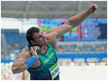 Imagem: Fotografia. Um homem robusto usando uniforme. Ele está segurando com a mão direita uma bola próxima ao rosto e o braço esquerdo estendido para frente.  Fim da imagem.
