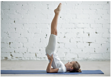 Imagem: Fotografia. Uma menina com camiseta e short branco. Ela está deitada com as costas sobre um colchonete e as pernas estendidas para cima com as mãos sobre o quadril. Fim da imagem.