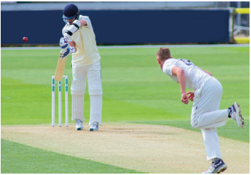 Imagem: Fotografia. Um homem usando uniforme branco e capacete, segurando um bastão chapado, atrás, uma bola. À direita, um homem com uniforme branco inclinado.  Fim da imagem.