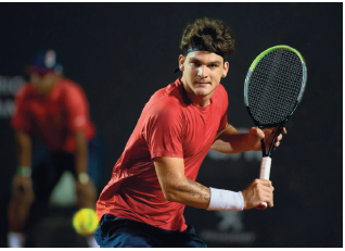 Imagem: Fotografia. Um homem com camiseta vermelha segurando uma raquete com a mão esquerda na direção de uma bola.  Fim da imagem.