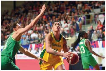 Imagem: Fotografia. À esquerda, uma mulher com uniforme verde. Ela está de perfil com os braços estendidos para cima. Ao lado, uma mulher com uniforme amarelo, segurando uma bola de basquete com as mãos olhando para cima. Atrás, outra mulher com uniforme verde. No fundo, arquibancada com diversas pessoas.  Fim da imagem.