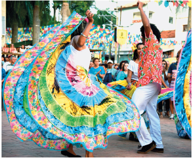 Imagem: Fotografia. Uma mulher com uma saia rodada colorida e rodada. Ao lado, um homem com camisa estampada e calça branca. Eles estão dançando em uma rua. Acima, várias bandeirinhas. No fundo, pessoas tocando instrumentos.  Fim da imagem.