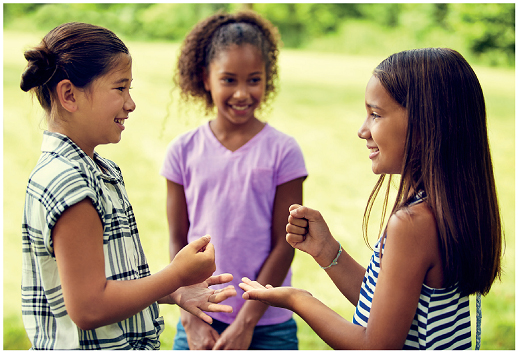 Imagem: Fotografia. Uma menina com cabelos presos, usando uma blusa quadriculada. Ela está de perfil com a mão esquerda fechada e a palma da mão direita aberta. Ao lado, uma menina com cabelos lisos na altura do ombro. Ela também está de perfil com a mão direita fechada e a palma da mão esquerda aberta. Atrás uma menina de cabelos presos, usando blusa roxa, olhando na direção das meninas e sorrindo.  Fim da imagem.