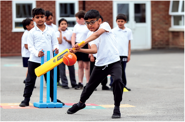 Imagem: Fotografia. Um menino uniformizado, segurando um bastão chapado na direção de uma bola. Atrás, um tripé azul e outro menino uniformizado segurando o tripé. No fundo, uma fileira de crianças uniformizadas.  Fim da imagem.