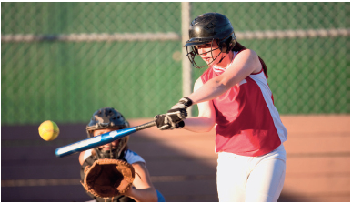 Imagem: Fotografia. Uma menina usando regata vermelha e um capacete segurando um bastão na direção de uma bola. Atrás, outra menina agachada com capacete com o braço estendido para frente com uma luva na mão.  Fim da imagem.