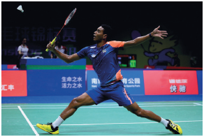 Imagem: Fotografia. Um homem uniformizado, segurando uma raquete na direção de uma peteca branca.  Fim da imagem.
