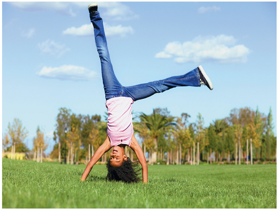 Imagem: Fotografia. Uma menina com cabelos crespos, usando uma blusa de alça e uma calça jeans. Ela está com as mãos no chão e as pernas para cima. Atrás, árvores.  Fim da imagem.