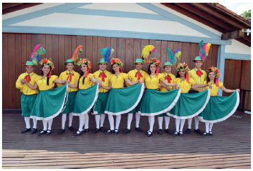 Imagem: Fotografia. Um grupo de pessoas uniformizadas. Homens, camisa amarela, short verde, chapéu com pernas. Mulheres, blusa amarela, saia verde e flores no cabelo. Os homens seguram a mão e as costas das mulheres. As mulheres, seguram na saia com a mão direita e sorriem.  Fim da imagem.