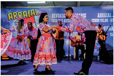 Imagem: Fotografia. Uma mulher usando um vestido roxo estampado. Ao lado, um homem usando uma camisa na mesma estampa do vestido e calça preta. Ele está inclinado segurando a mão da mulher. Atrás, outro casal na mesma posição deles. No fundo, homens segurando instrumentos de corda. Fim da imagem.