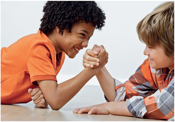 Imagem: Fotografia. Um menino com cabelos cacheados, usando uma camisa vermelha. Ele está de perfil segurando a mão de um menino com cabelos loiros, usando blusa xadrez. Eles estão com os cotovelos sobre a mesa.  Fim da imagem.