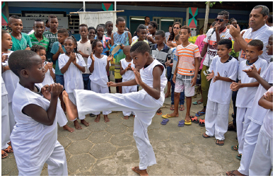 Imagem: Fotografia. À esquerda, um menino negro usando roupas brancas, com os braços flexionados e as mãos fechadas próximo ao rosto. Ao lado, outro menino negro, com roupas brancas com a perna direita estendida para frente na direção do menino. Ao redor, um círculo com pessoas e crianças batendo palmas  Fim da imagem.