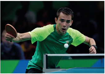 Imagem: Fotografia. Um homem com cabelos curtos, segurando uma raquete com a mão esquerda, olhando na direção de uma bola, que passa por cima de uma rede em cima de uma mesa.  Fim da imagem.