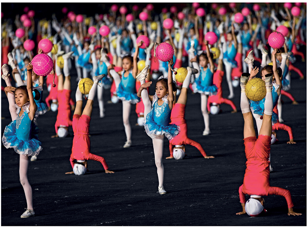Imagem: Fotografia. Um grupo de meninas com vestido azul e meia calça. Elas estão com a perna esquerda estendida para cima com o braço esquerdo ao redor da perna, a mão direita estendida para cima segurando uma bola, e o pé direito no chão. Entre elas, um grupo de meninas com a cabeça e as mãos no chão, as pernas estendidas para cima, segurando uma bola entre os tornozelos.  Fim da imagem.