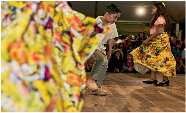 Imagem: Fotografia. Um menino usando camisa, calça e um tamanco. Ao lado, uma menina, usando blusa e saia de chita, e sapatos de saltos. Eles estão de pé um de frente para outro em um palco. No fundo, pessoas sentadas, olhando para eles.  Fim da imagem.