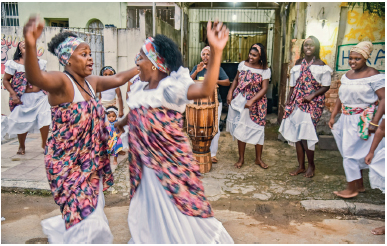 Imagem: Fotografia. Duas mulheres com vestido branco e um tecido colorido em volta do corpo e um lenço no cabelo. Elas estão de frente uma para outra, com as mãos estendidas para cima. Atrás, outras mulheres em uma roda.  Fim da imagem.