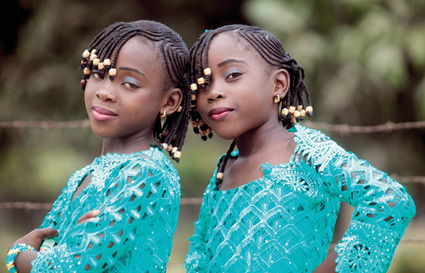 Imagem: Fotografia. Duas meninas com tranças, maquiagem e roupa azul estão sorrindo.  Fim da imagem.