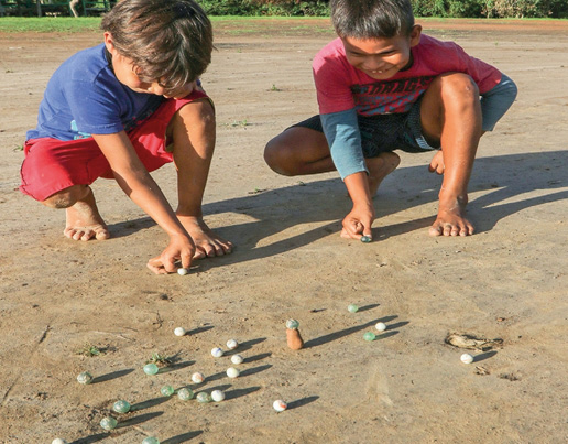 Imagem: Fotografia. Dois meninos estão descalços, agachados e segurando bolinhas de gude. Na frente deles há várias bolinhas espalhadas no chão. Fim da imagem.