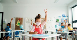 Imagem: Fotografia. Crianças com máscaras estão sentadas em carteiras escolares e com as mãos levantadas.  Fim da imagem.