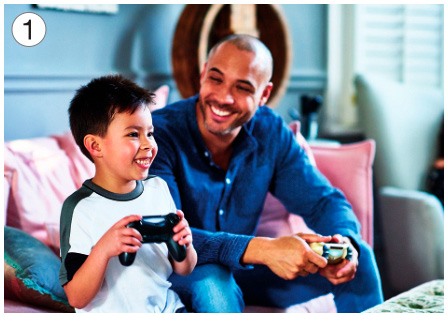 Imagem: Fotografia 1. Um menino está sorrindo, sentado em um sofá e segurando um controle de videogame. Ao seu lado, um homem está sentado no sofá, o observando e sorrindo. Fim da imagem.