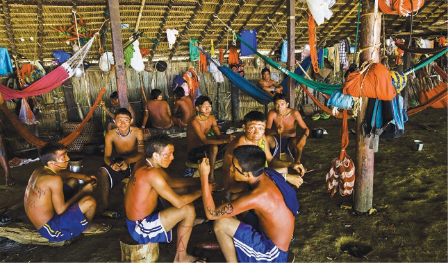 Imagem: Fotografia. Vários homens indígenas com pinturas no rosto, sem camisa e com bermuda azul estão sentados dentro de uma oca grande. Em volta deles há várias redes coloridas penduradas. Fim da imagem.