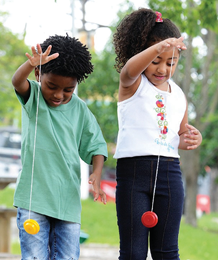 Imagem: Fotografia. Duas crianças estão sorrindo com as mãos para cima e com cordas de ioiôs presas nos dedos. Fim da imagem.