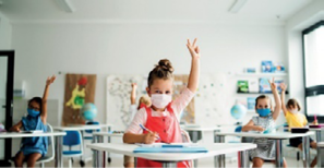 Imagem: Fotografia. Crianças com máscaras estão sentadas em carteiras escolares e com as mãos levantadas.  Fim da imagem.