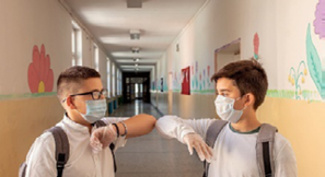 Imagem: Fotografia. Dois meninos com máscaras estão encostando os cotovelos um no outro. Ao fundo, portas em um corredor. Fim da imagem.