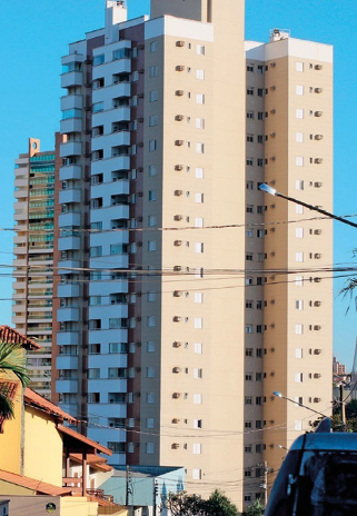 Imagem: Fotografia. Um prédio alto com paredes bege. Em volta há mais prédios e casas. Fim da imagem.