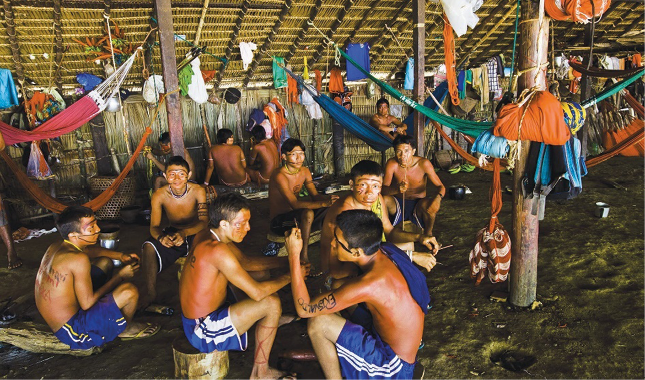 Imagem: Fotografia. Vários homens indígenas com pinturas no rosto, sem camisa e com bermuda azul estão sentados dentro de uma oca grande. Em volta deles há várias redes coloridas penduradas. Fim da imagem.