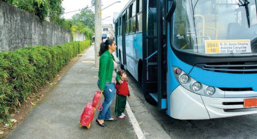 Imagem: Fotografia. Uma mulher está segurando a mão de um menino e com a outra mão, ela segura uma mochila de rodinhas. Na frente deles há um ônibus parado com a porta aberta.  Fim da imagem.