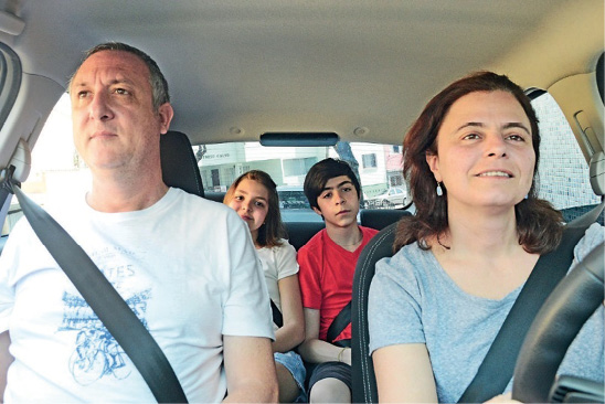 Imagem: Fotografia. Um casal está sentado nos bancos da frente de um carro e duas crianças estão sentadas nos bancos traseiros. Todos estão com cinto de segurando em volta do corpo. Fim da imagem.