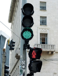 Imagem: Fotografia. Na parte superior, um semáforo com a luz verde acesa. Na parte inferior, um semáforo de pedestres com a luz vermelha acesa.  Fim da imagem.
