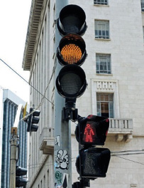 Imagem: Fotografia. Na parte superior, um semáforo com a luz amarela acesa. Na parte inferior, um semáforo de pedestres com a luz vermelha acesa.  Fim da imagem.